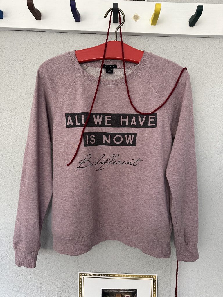 Pullover mit Spruch Rosa Pullover an einer Wand mit dem Spruch "All we have is now"